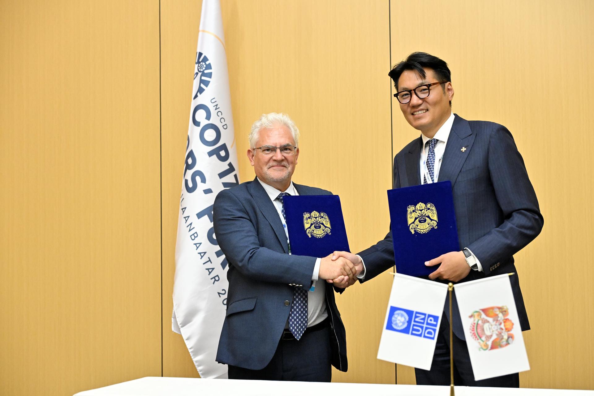 Two men in suits shake hands during a signing ceremony, each holding a blue folder with a crest.