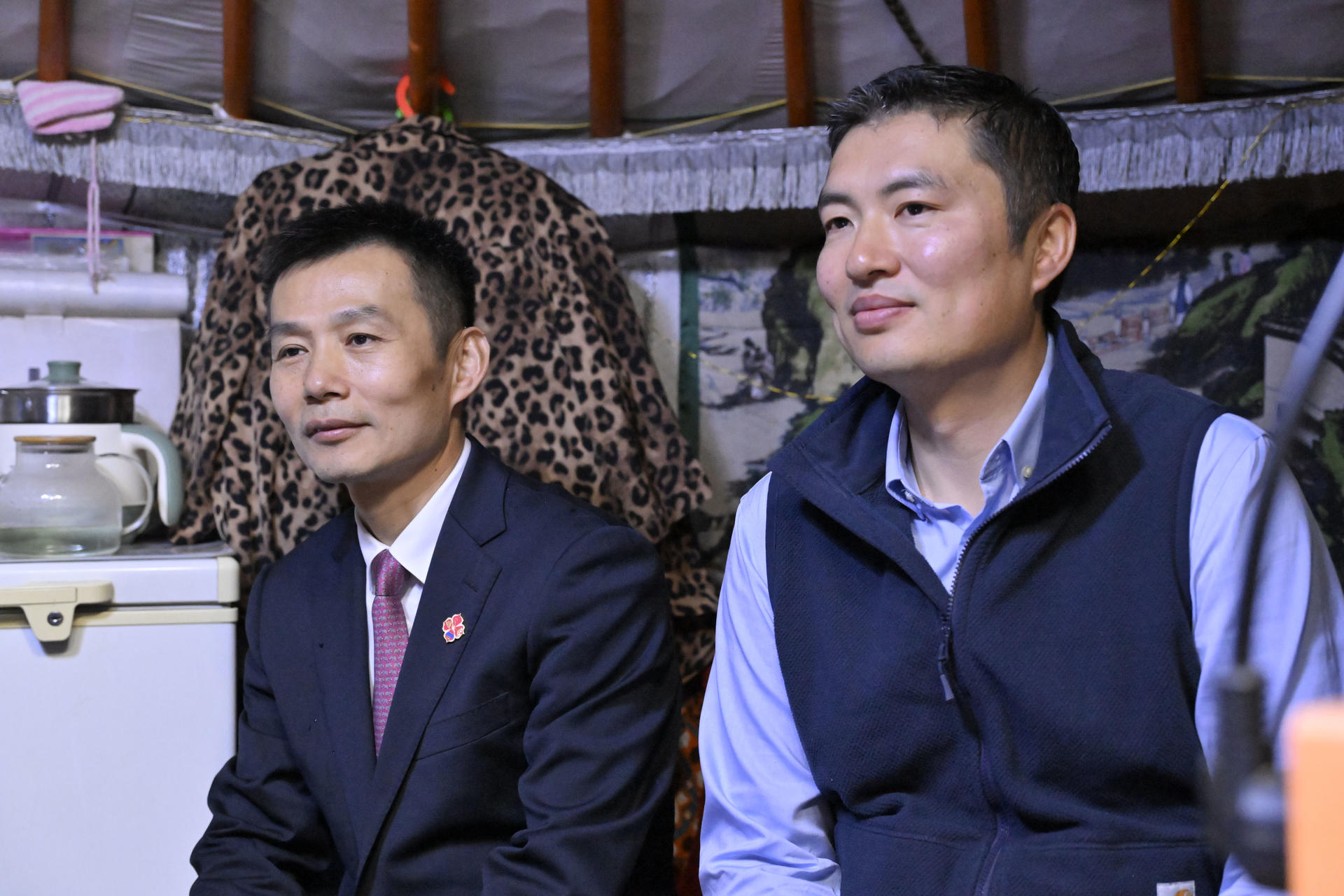 Two men sit indoors side by side; one in a suit, the other in a blue vest, patterned fabric behind.