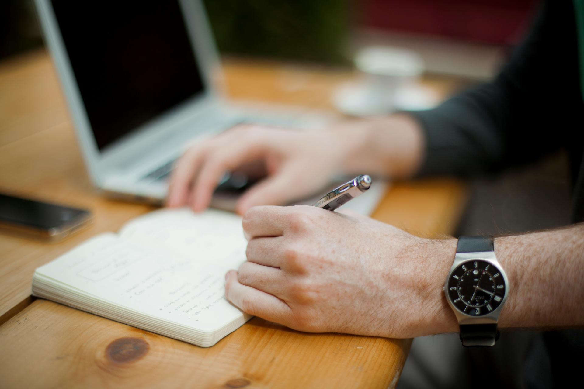 Close-up of hands with a pen writing in a notebook on a wooden desk, laptop nearby.