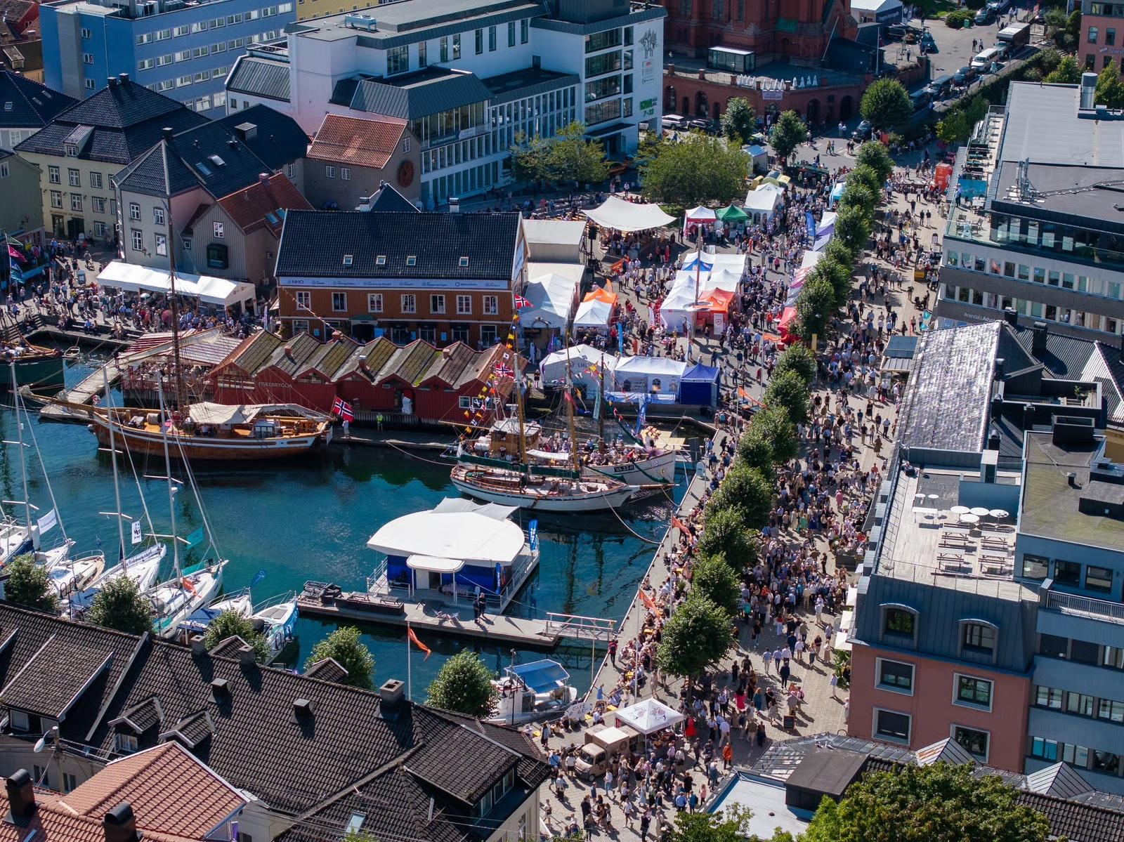 Aerial view of a harbor town with colorful buildings, boats, and a crowded waterfront promenade.
