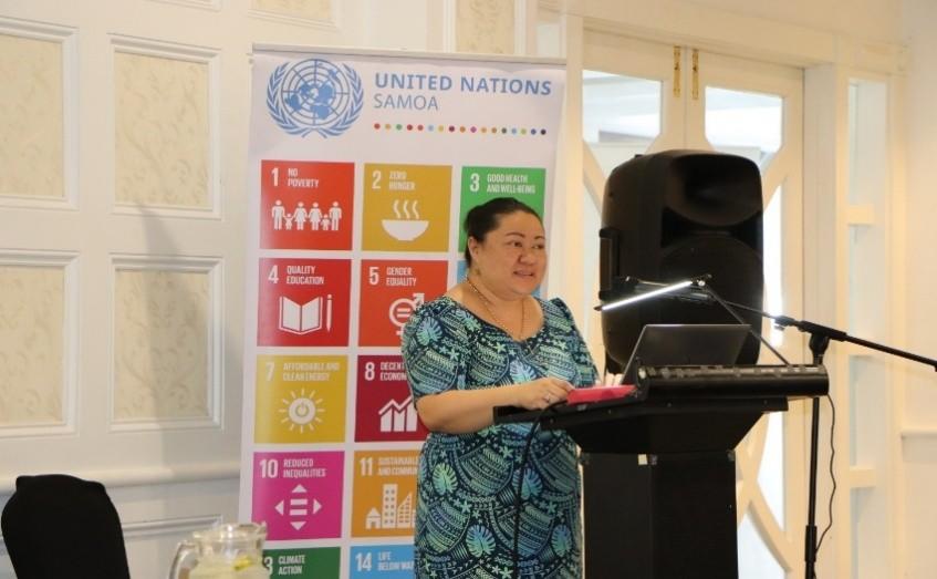 Photo: woman speaker at a podium with laptop; UN SDGs banner in a sunlit room.