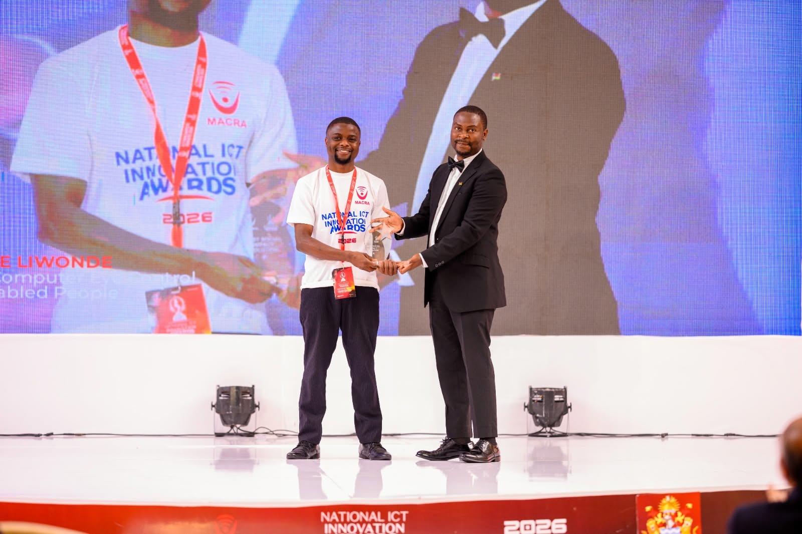 Onstage award ceremony: man in white T-shirt receives trophy from presenter, backdrop screen.