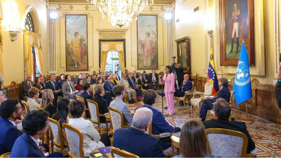 Elegant chandelier-lit hall; audience sits as a speaker addresses a podium, blue flag nearby.