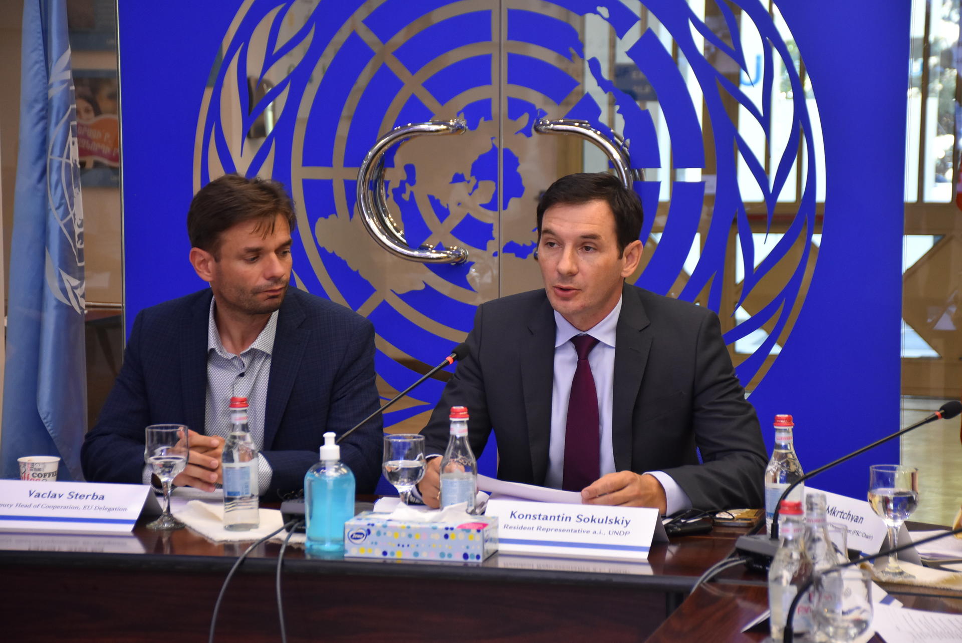 Photograph of two men at a conference table in front of a blue United Nations backdrop.