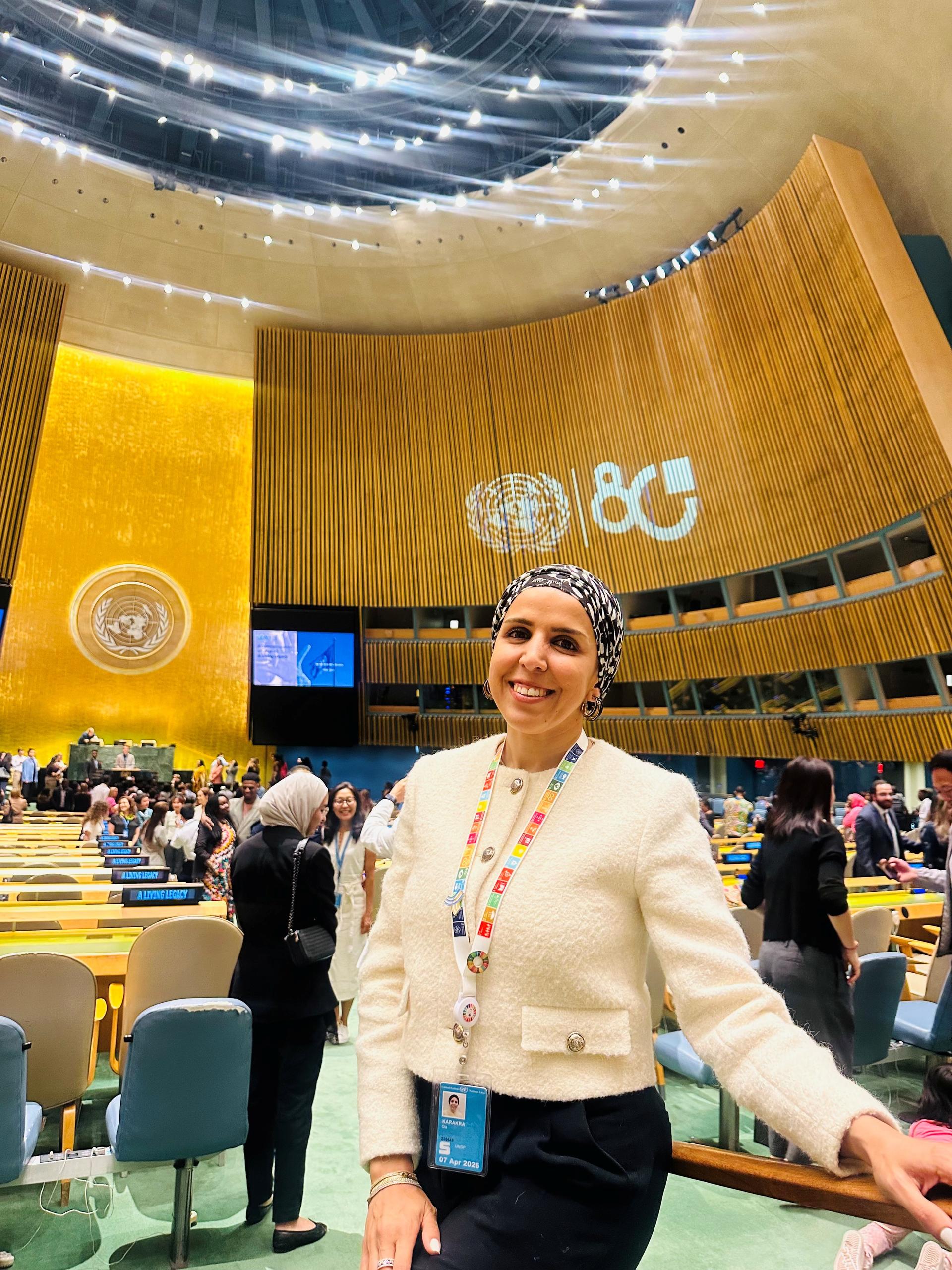 Smiling woman in a white top with a badge in a conference hall with wooden walls and attendees.