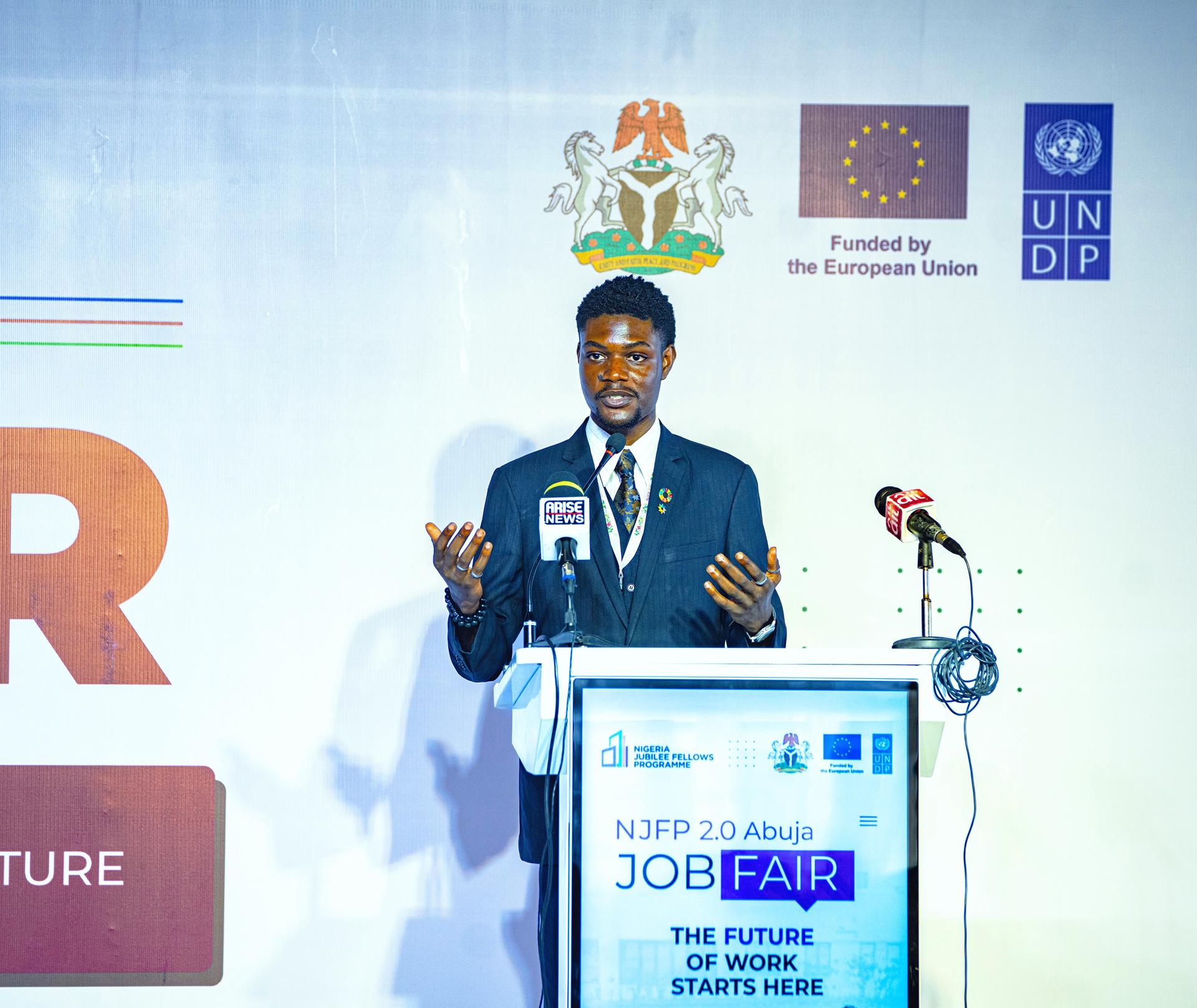 Photograph of a man in a suit giving a speech at a job fair stage with UNDP banners.