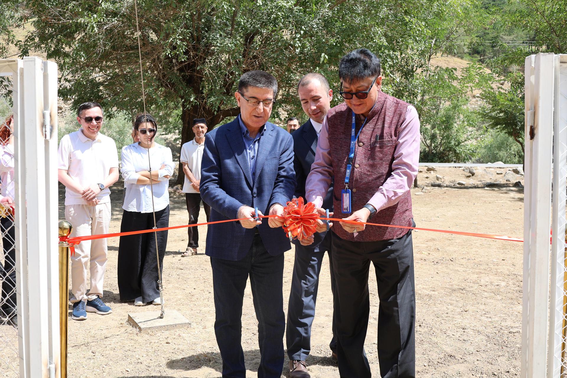 Photo: Two men cut a red ribbon at an outdoor ribbon-cutting ceremony, while onlookers watch.