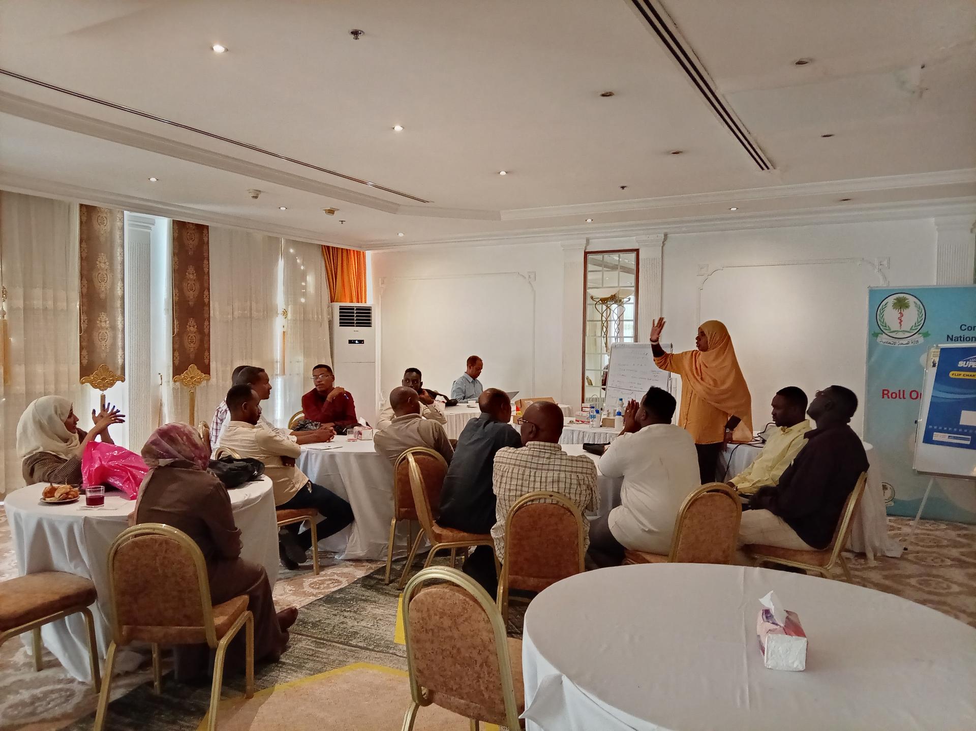 Seminar in a bright conference room with round tables; presenter at front near a screen.