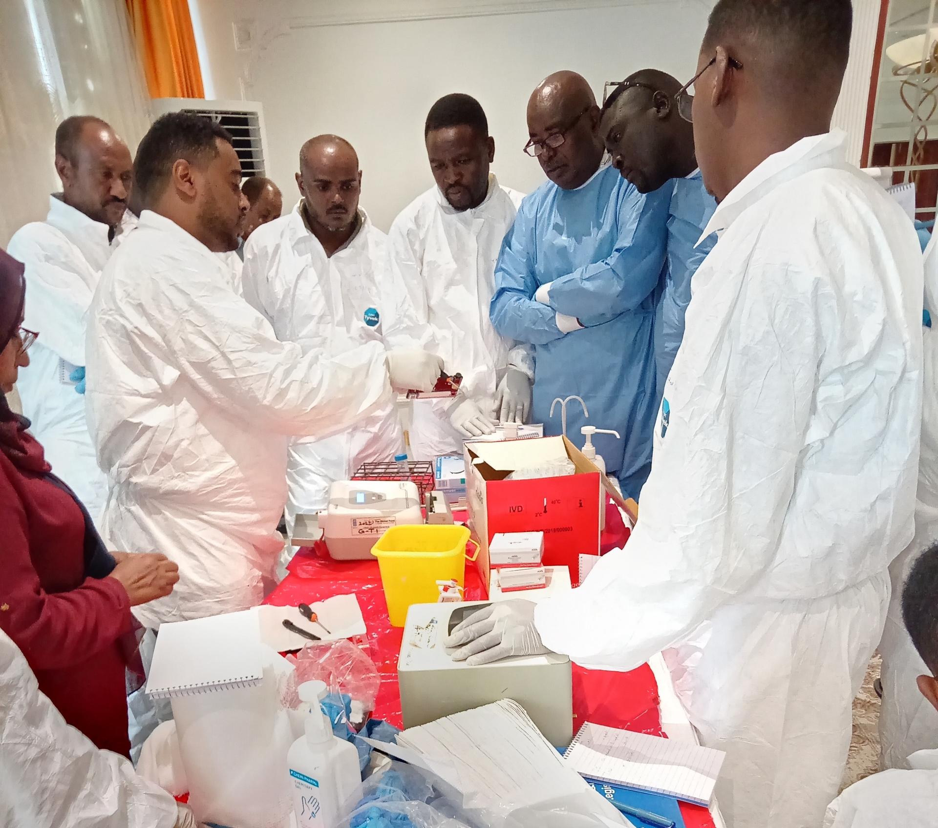 Group of people in white coats and blue protective suits around a table with medical supplies.