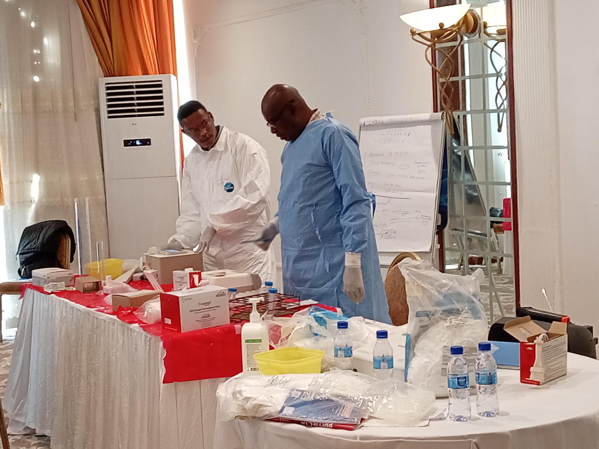 Photo: two researchers in lab coats stand by a long table with supplies in a clinical room.