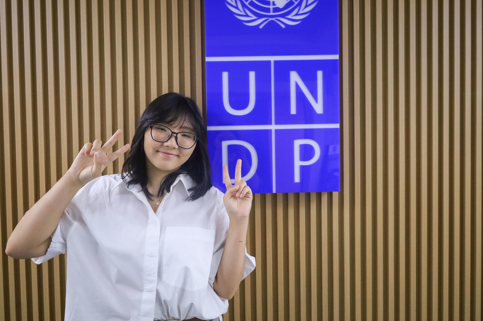 Smiling woman in a white blouse making peace signs in front of a UNDP sign.