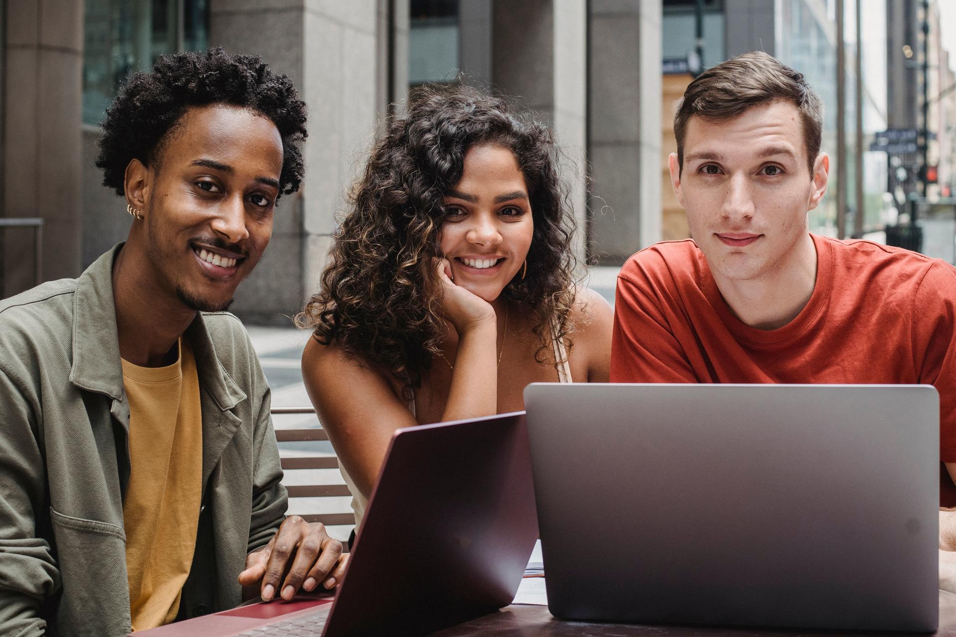 Three friends sit at a sidewalk table outdoors with laptops, smiling at the camera.
