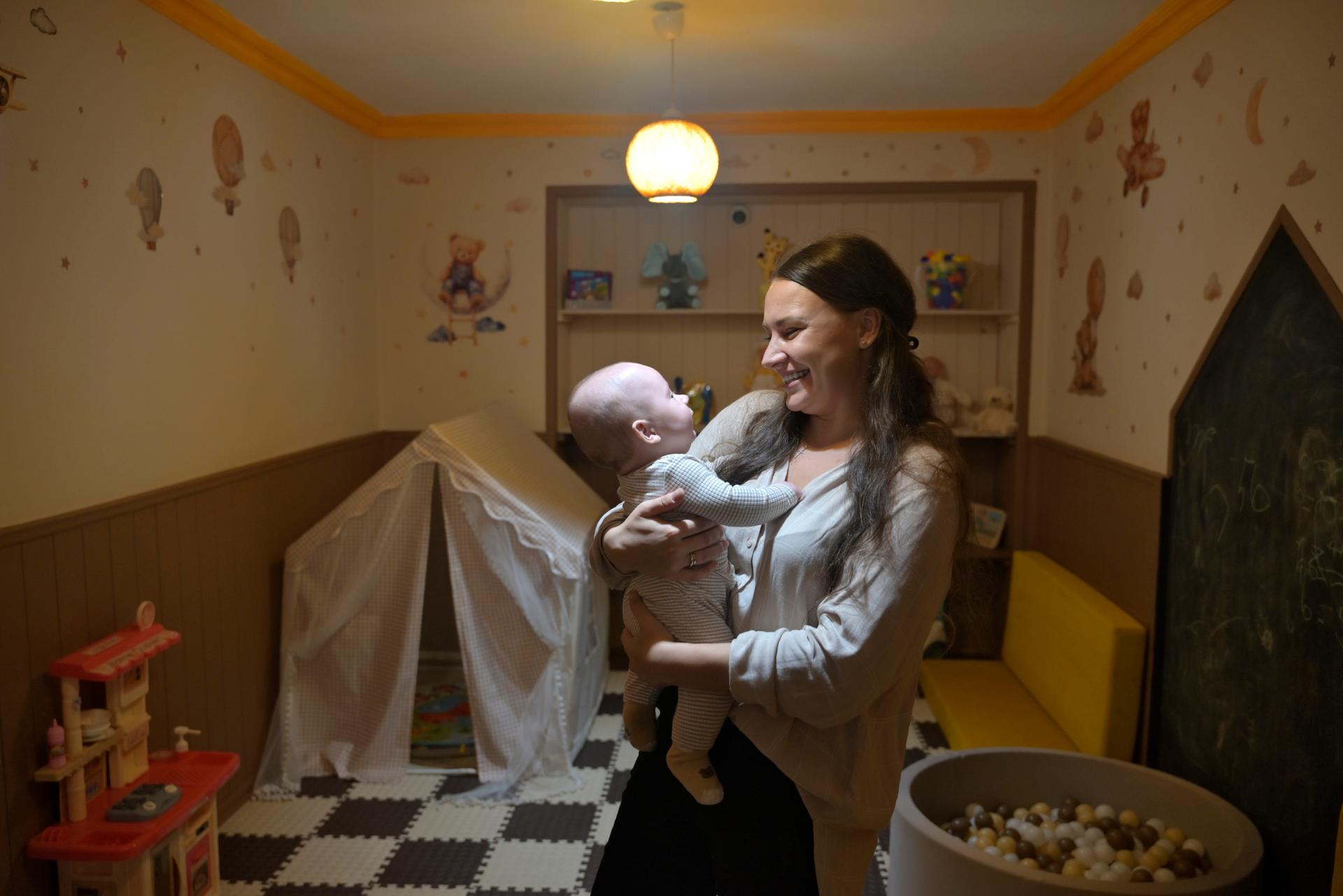 Young woman holding a baby in a warm, cozy room with a black-and-white checkered floor.