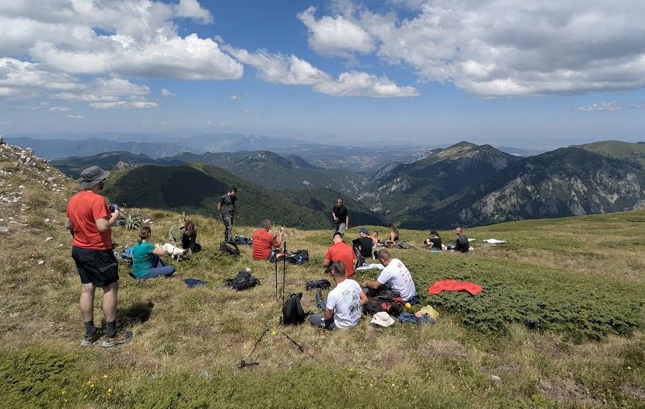 Photograph: Hikers resting on a grassy hillside with backpacks, distant mountains and blue sky.