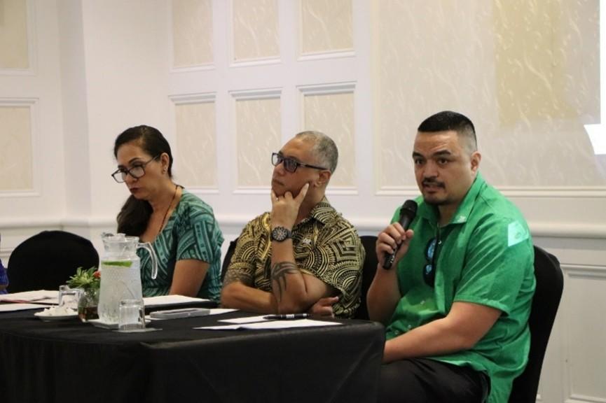 Three panelists sit at a conference table; man in camouflage speaks into a microphone.