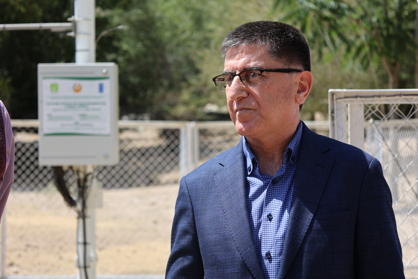 Photograph of a man in a blue suit outdoors, wearing glasses, with a fence and sign behind.