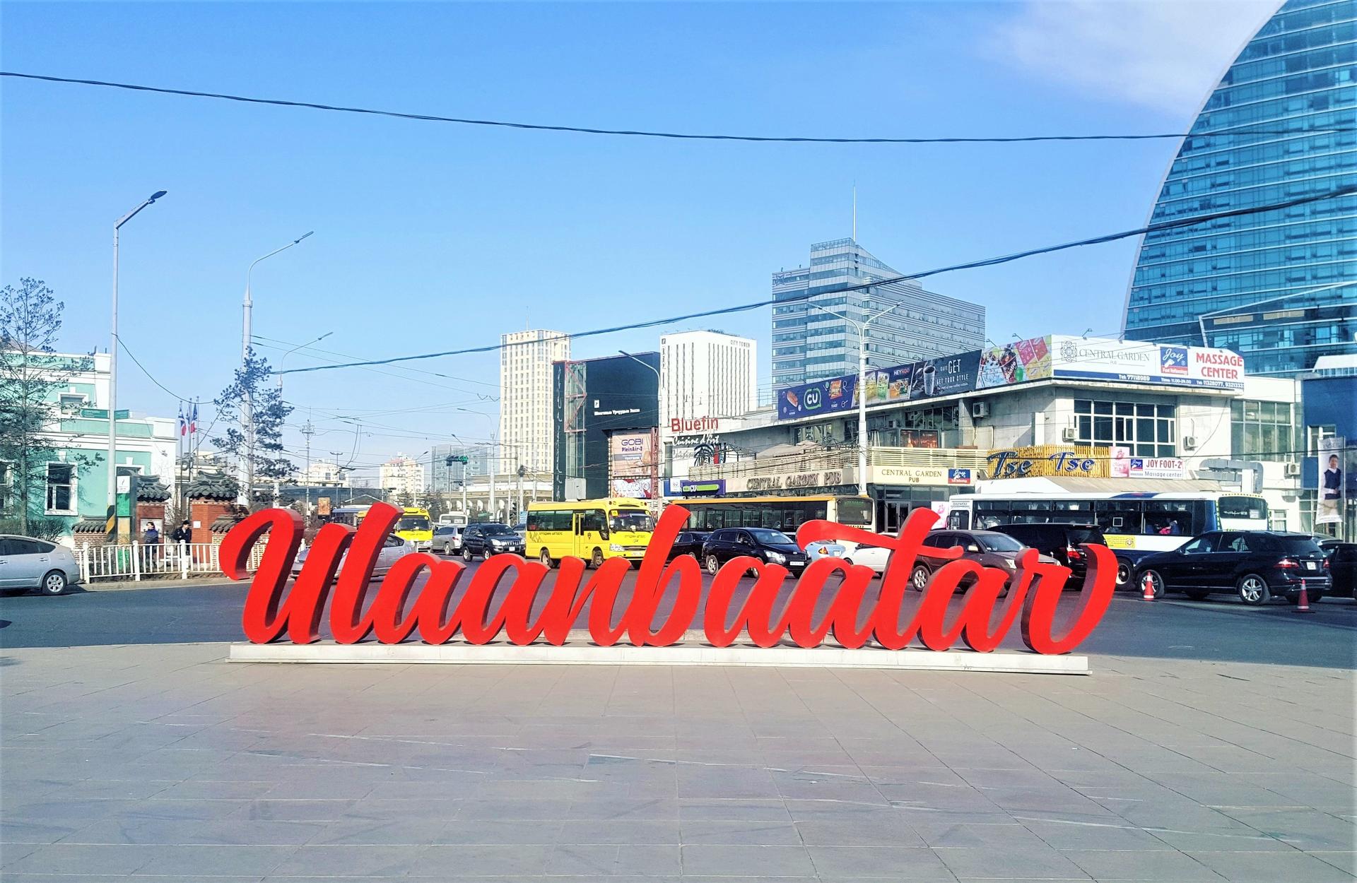 Red Ulaanbaatar sign sculpture on a sunny street with buses and modern buildings in the background.