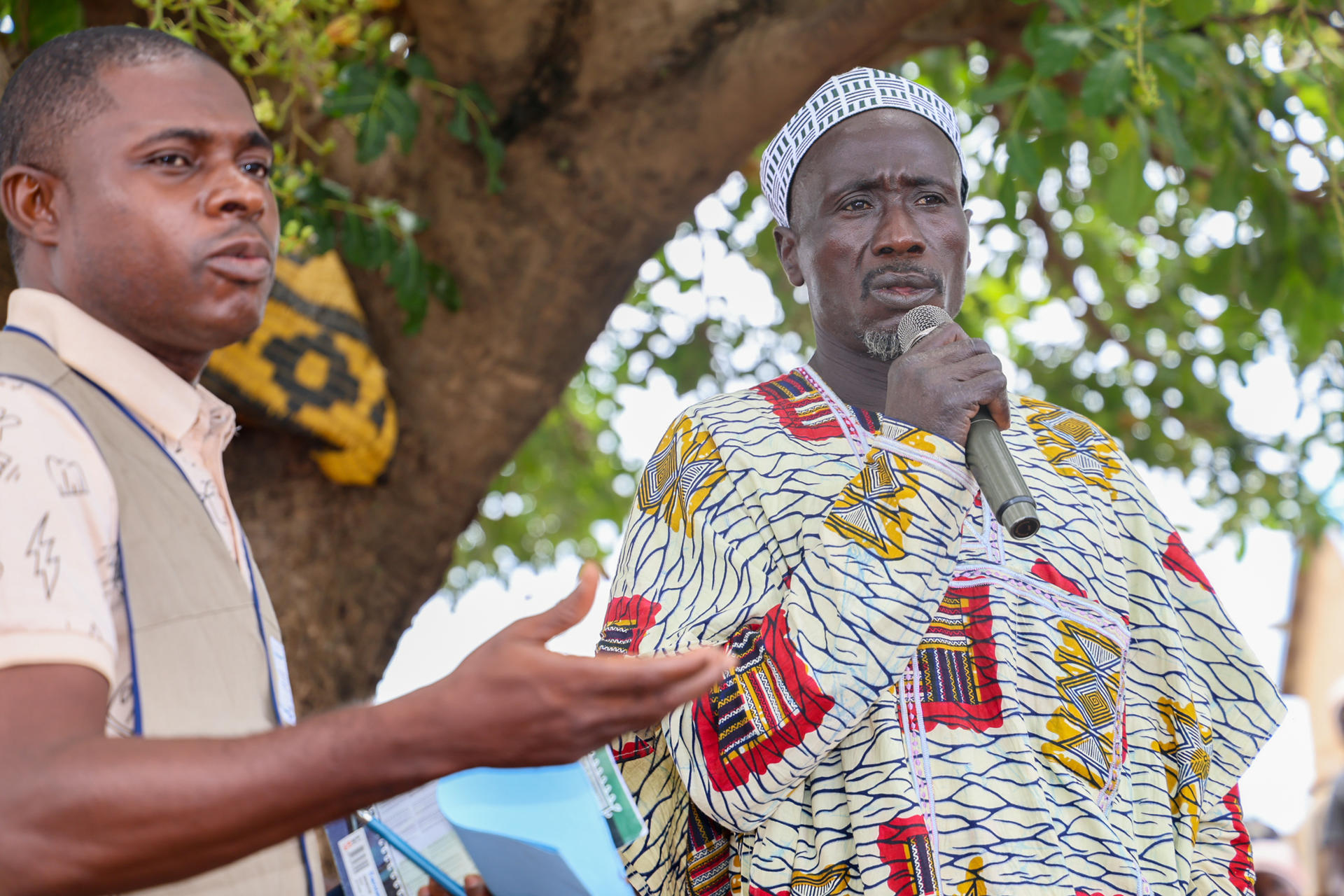 Two men outdoors under a tree; one in a patterned shirt and white cap, the other in beige, gesturing.