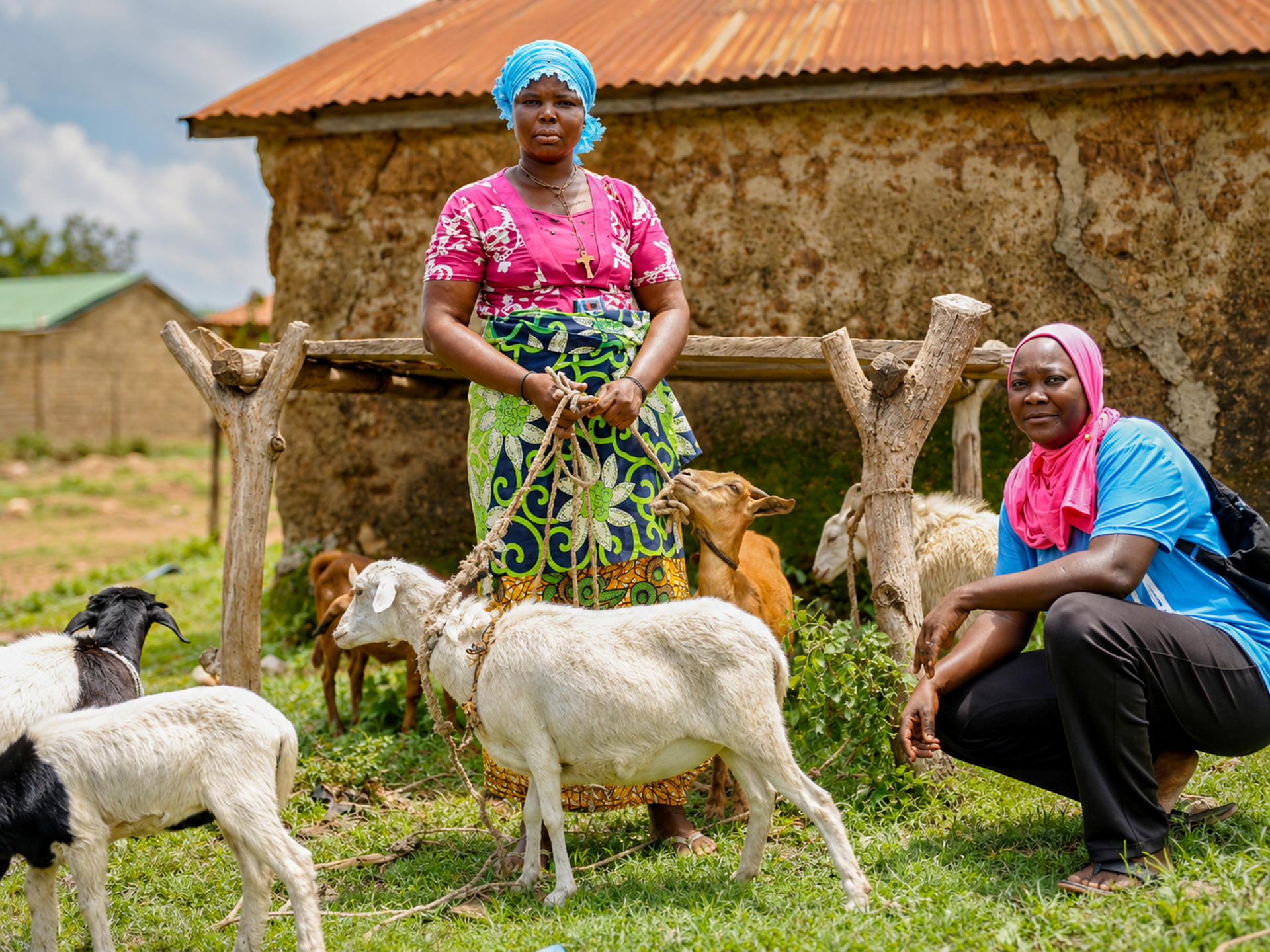 Two women with goats in a rural village yard; mud-brick huts and a wooden fence in back.