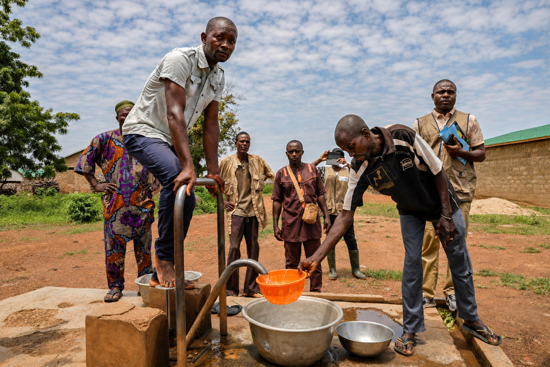 People gather around a hand pump in a rural village, filling an orange bucket.