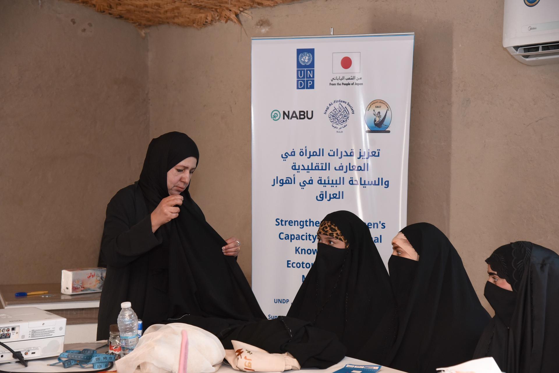 Women in black hijabs sit around a table as a speaker stands; banners with logos behind.