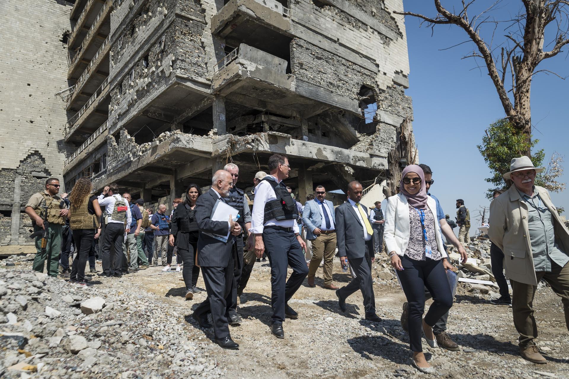 Group of people in business attire walking through rubble beside a partly destroyed building.