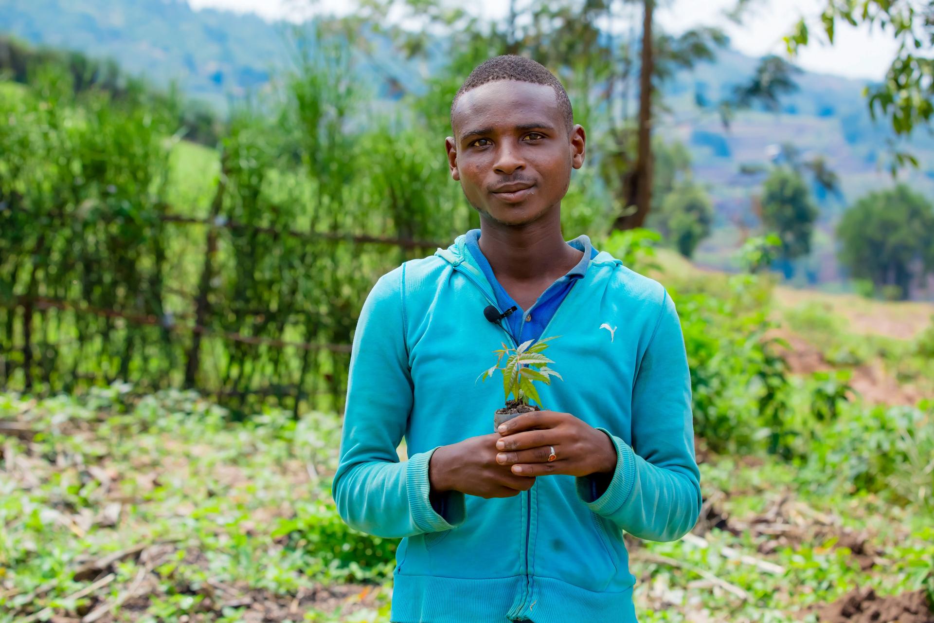 Photograph of a young man in a teal shirt holding a small plant in a green field.