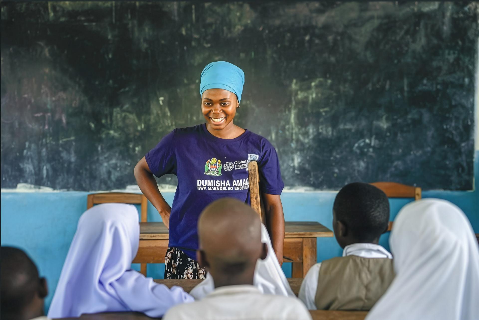 Female instructor in blue hijab and purple shirt addressing seated children in a classroom.