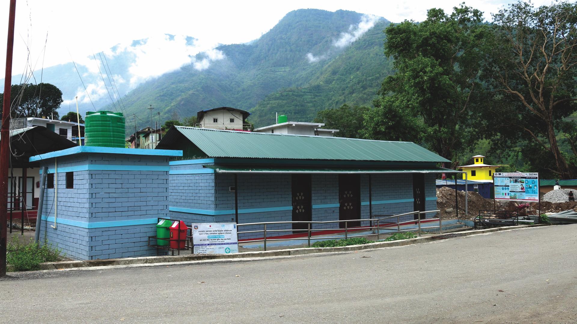 Photograph of blue storefronts along a dusty street with green mountains in the distance.