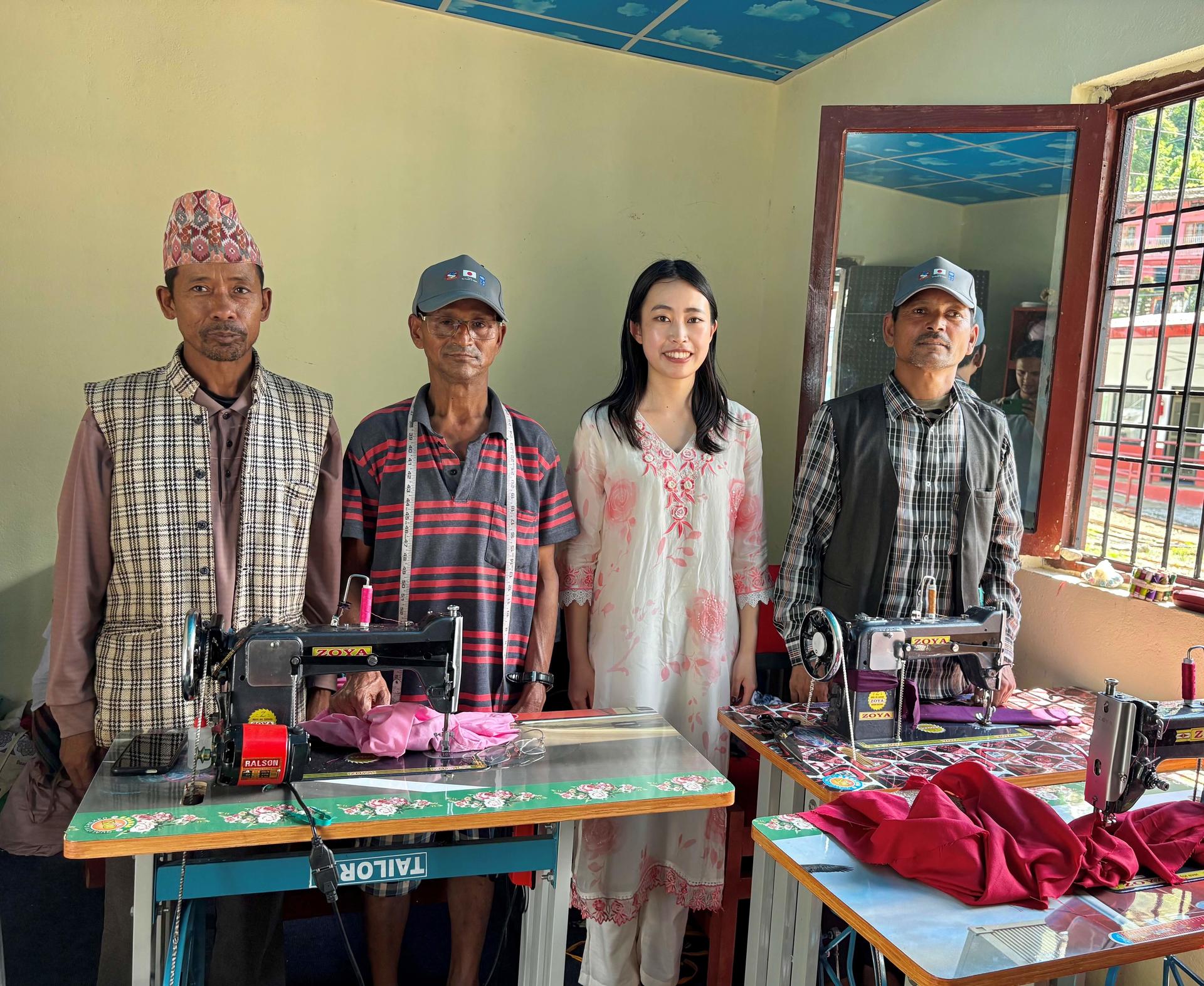 Four individuals stand behind sewing tables with machines and fabric in a bright room.