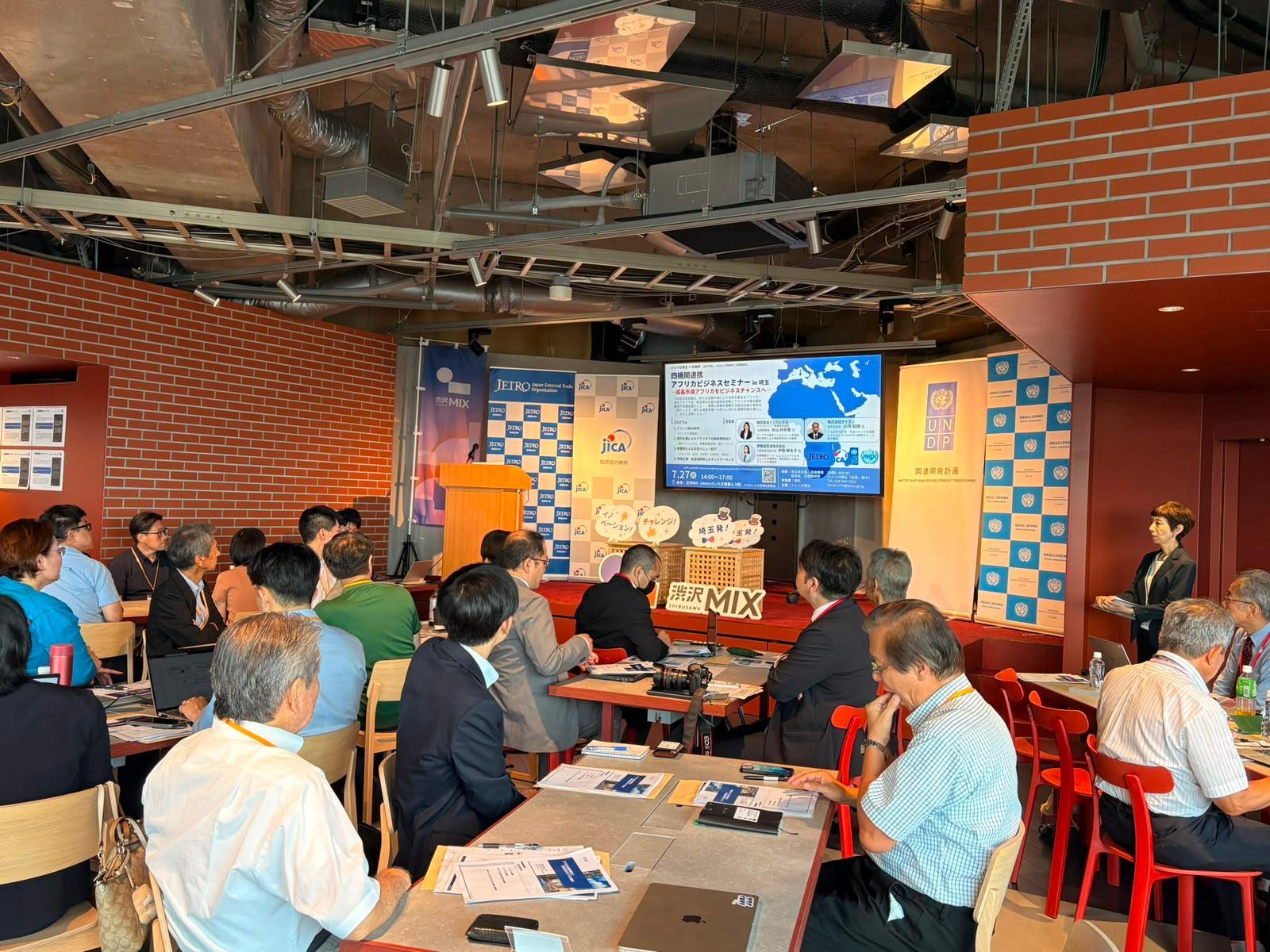 People seated around tables in a conference room, presentation screen at the front.