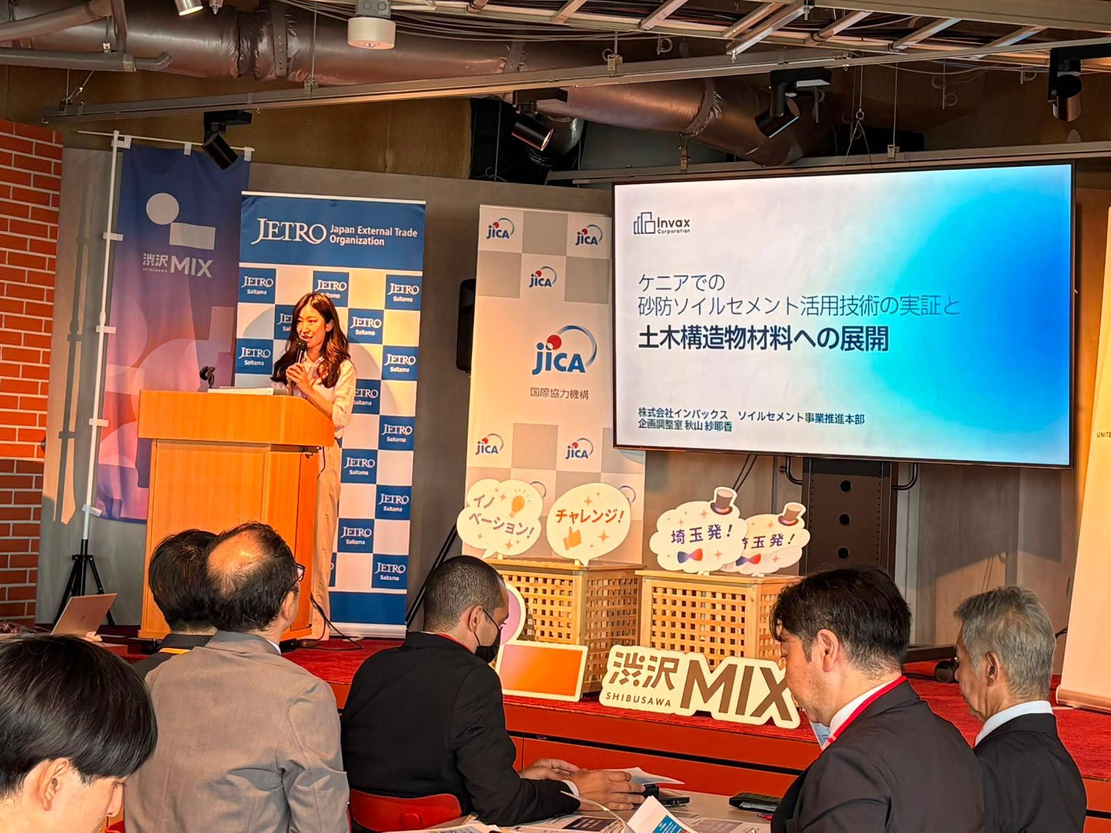 Conference stage with speaker at orange podium, banners with logos, blue presentation screen.