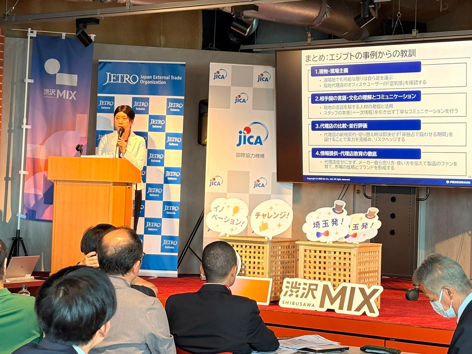 Photograph of a presenter at a podium addressing an audience in a conference hall with banners and a screen.