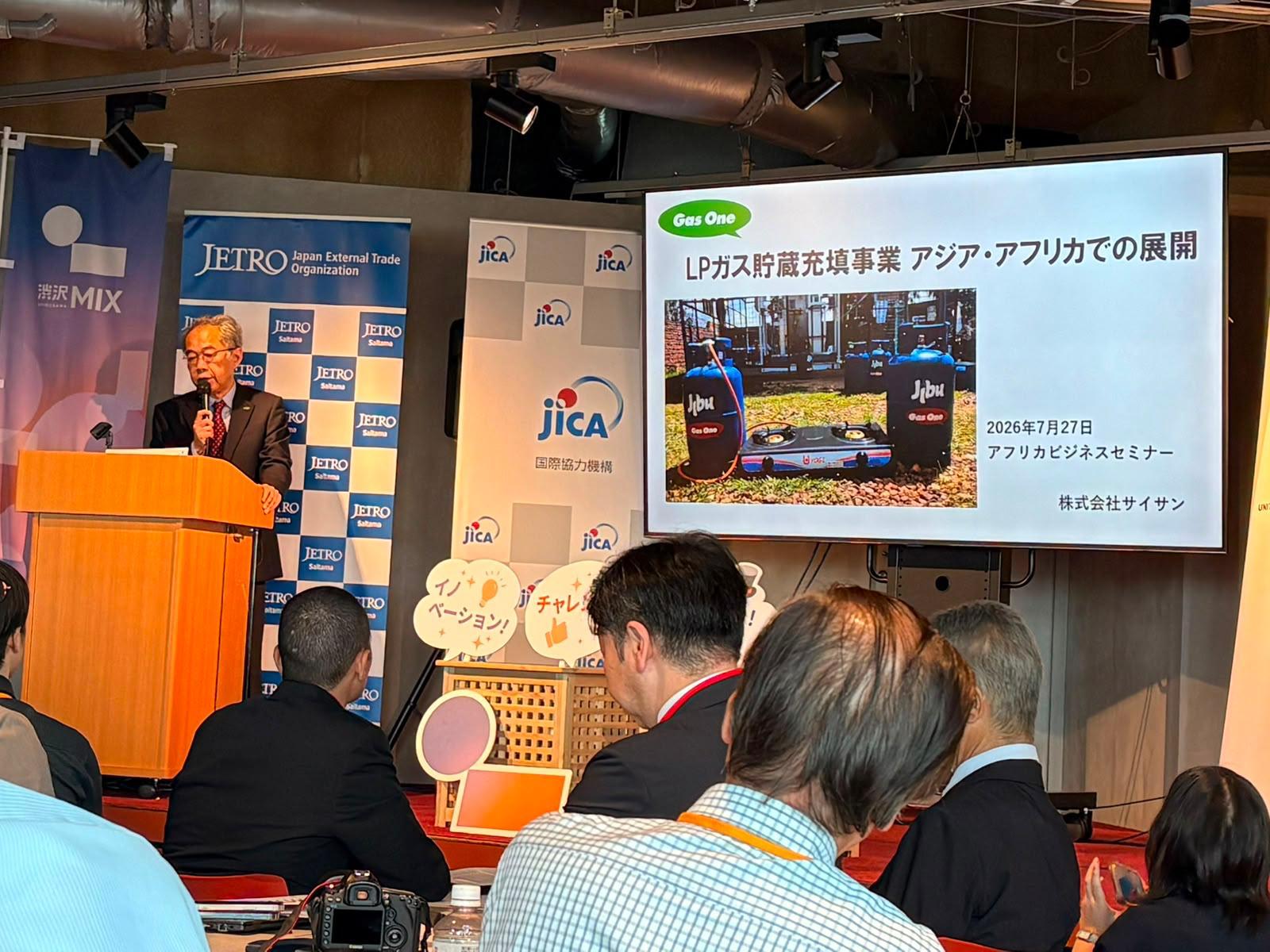Speaker at podium presenting to an audience in a conference room; slide in Japanese on screen with JICA banners on stage.