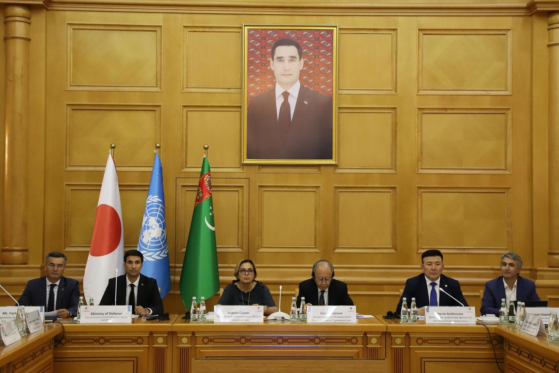 Officials seated at a long wooden table in a paneled room with flags and a framed portrait.