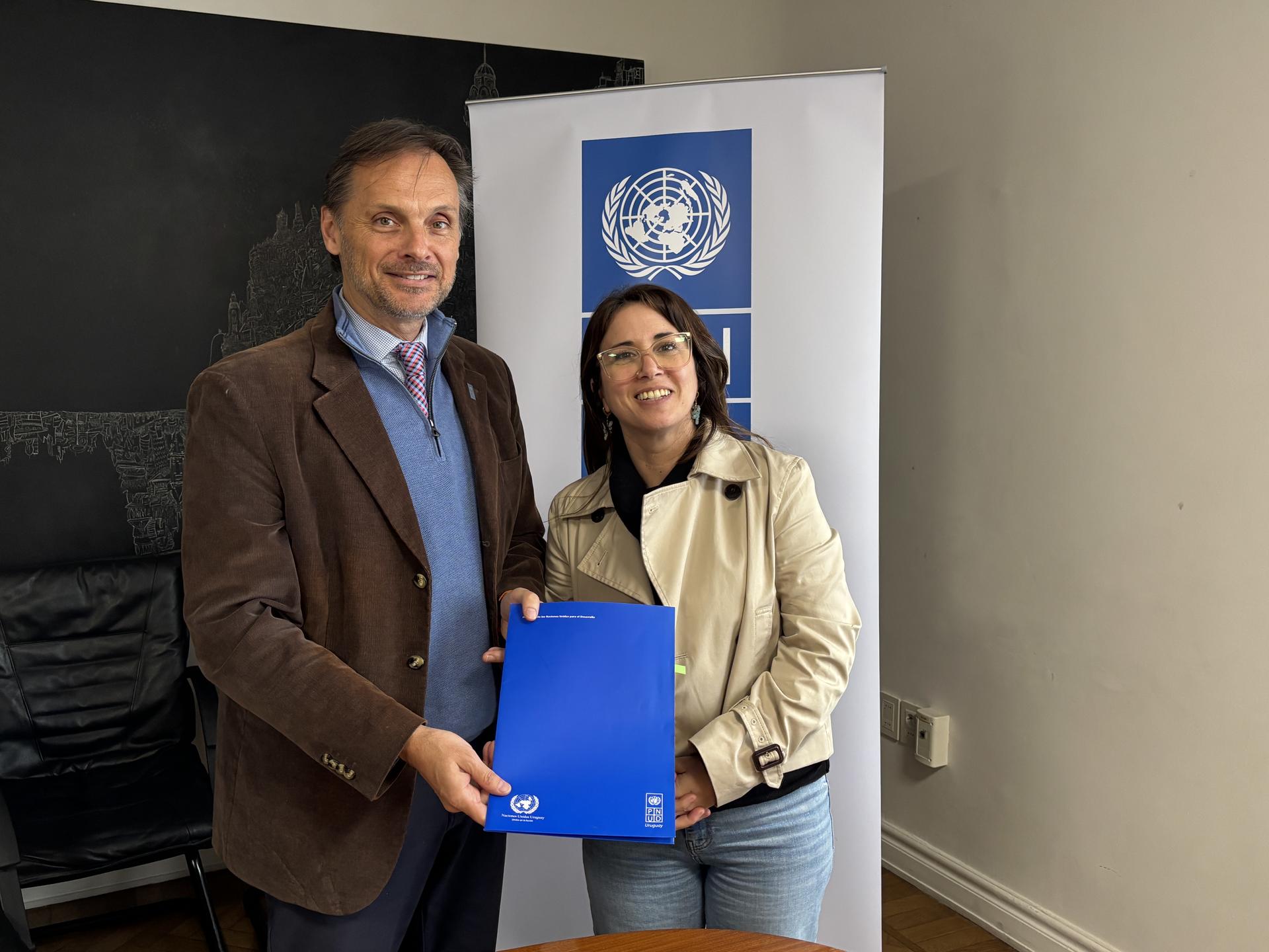 Two people indoors, smiling, holding a blue folder; banner with a globe logo behind.