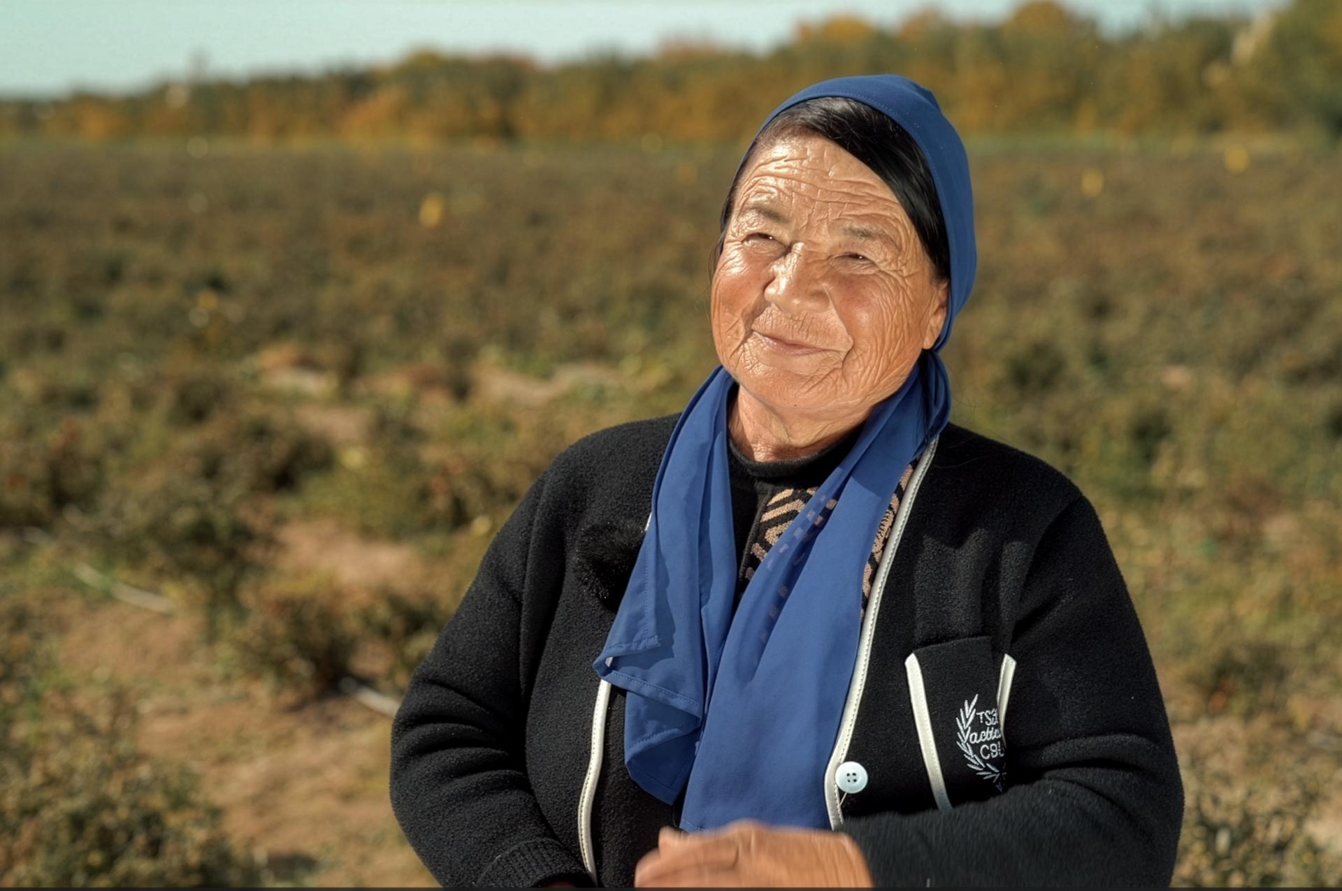 Smiling woman in a blue headscarf and black jacket in a dry field.