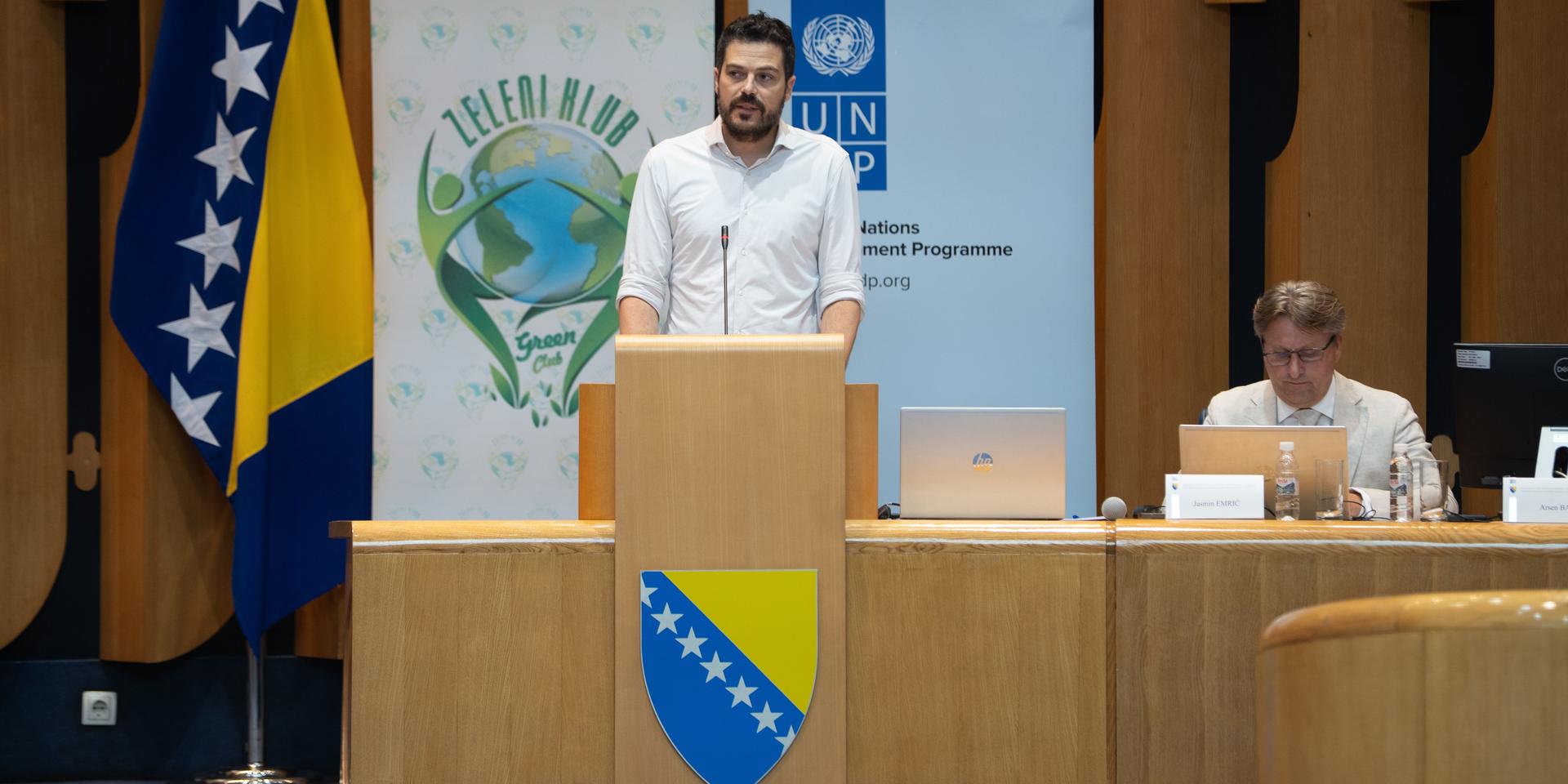 Man at podium speaking in a conference room; two officials seated at a desk, Bosnian flag visible.