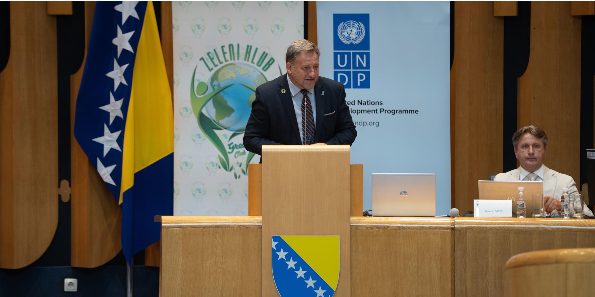 Photograph of a man speaking at a podium during a conference, with Bosnian flags in the background.