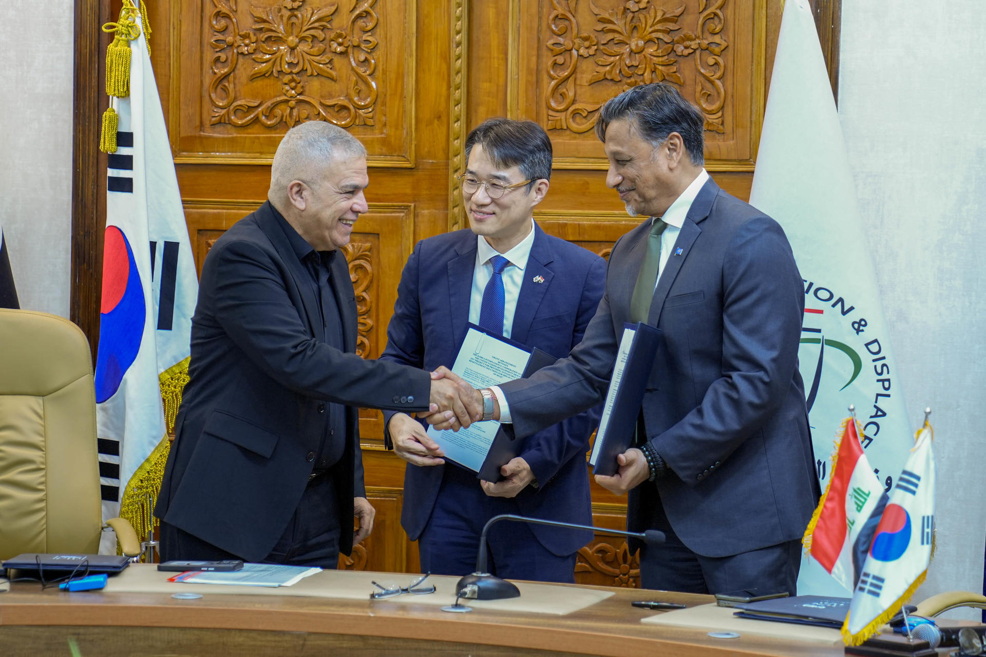 Three men in suits shake hands at a formal meeting with flags on the table.