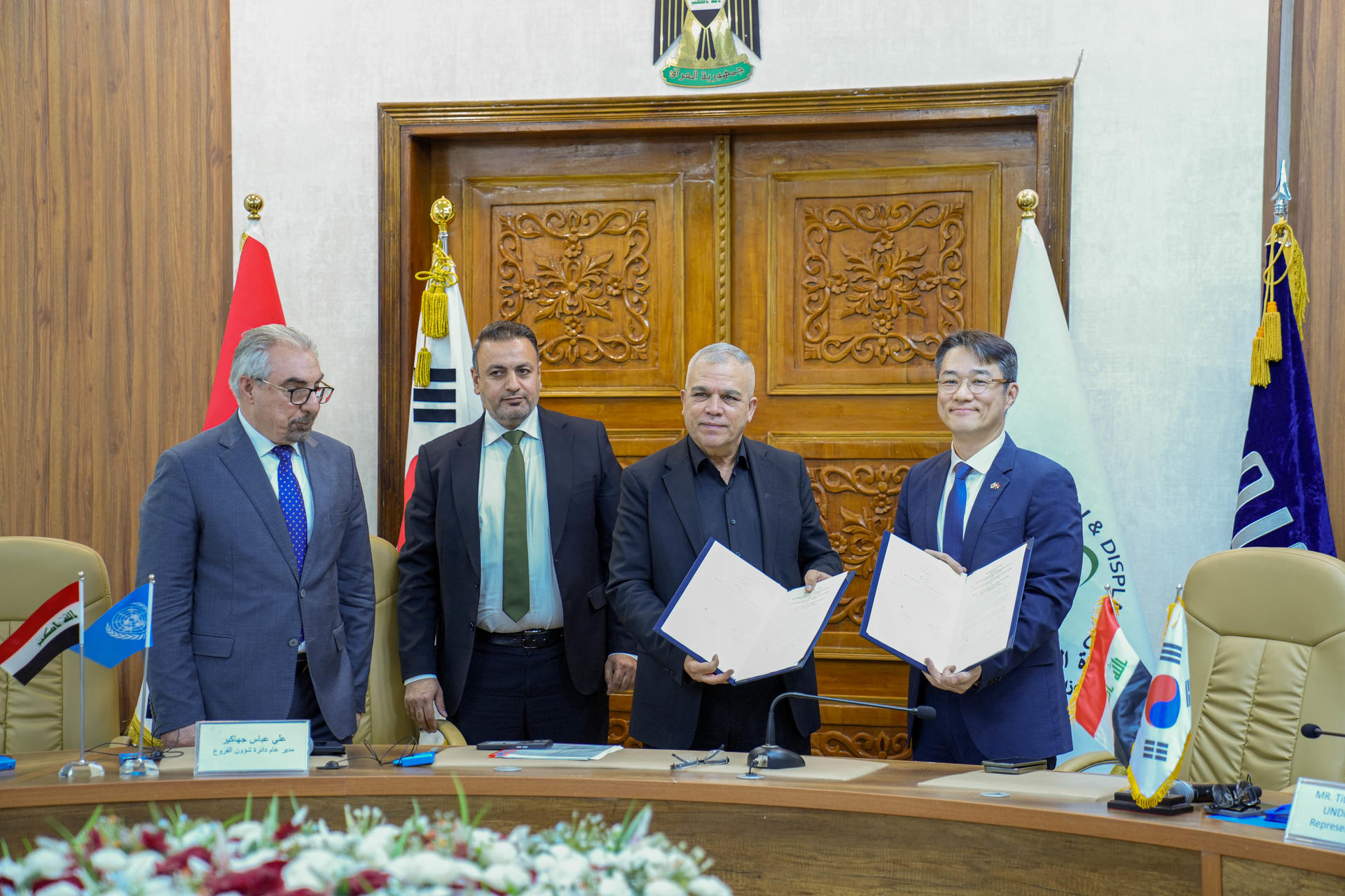 Photograph of five men in suits signing documents at a formal ceremony, with flags and a crest.