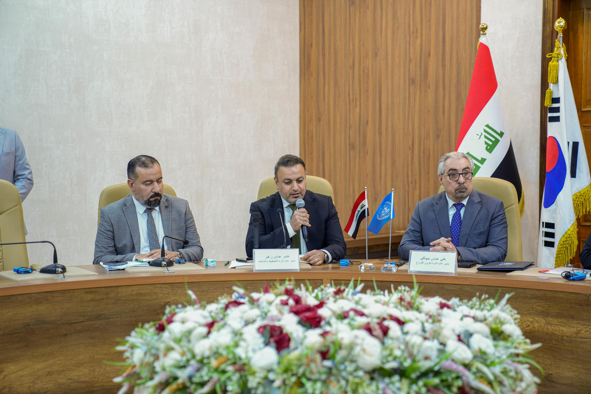 Three men in suits sit at a conference table with a floral centerpiece and flags in the background.