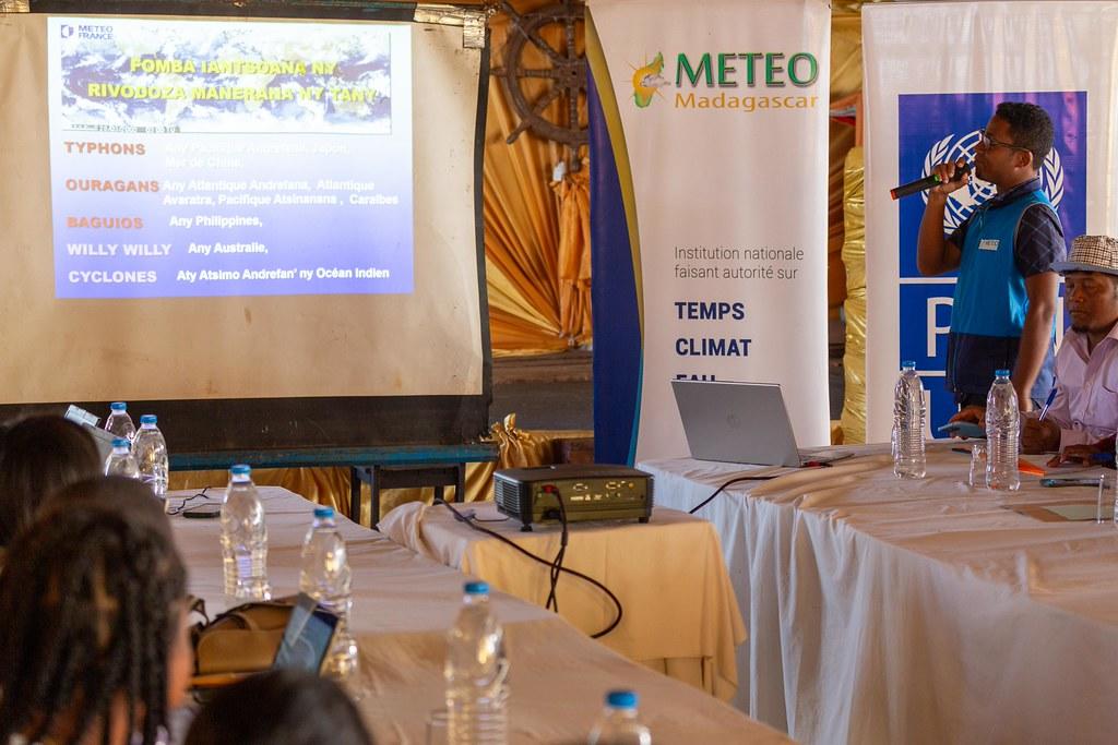 Presentation room; slide on projector screen, attendees seated at tables, banners in background.