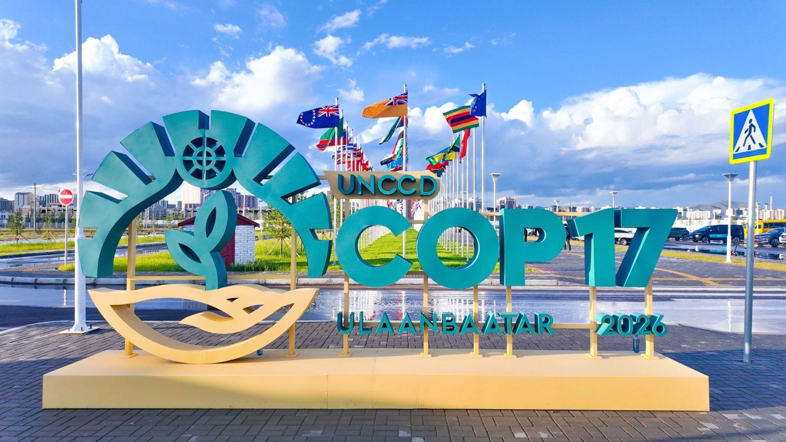 Colorful seaside sign sculpture with a wheel logo and teal letters on a yellow pedestal, against blue sky.