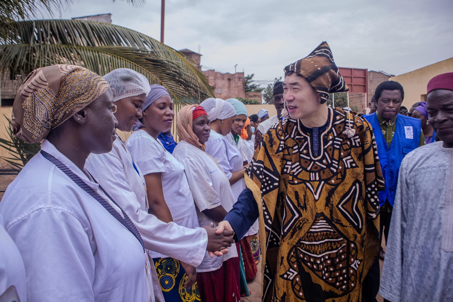 Group of villagers outdoors; white robes shake hands with a figure in bold patterned attire.