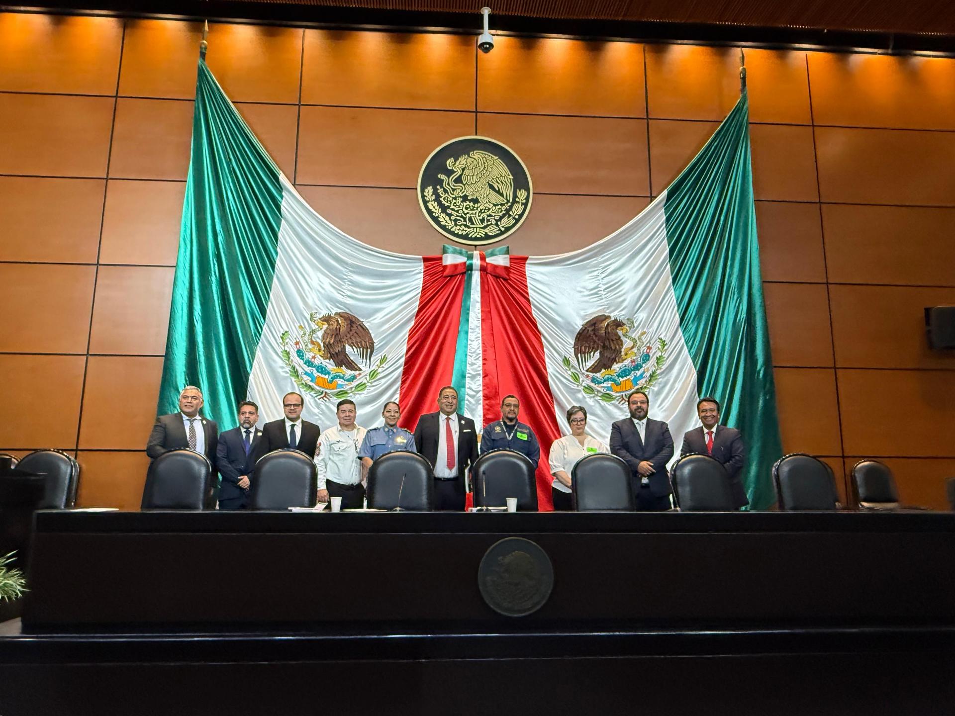 Panel of officials on a stage behind a long desk, framed by large Mexican flags.