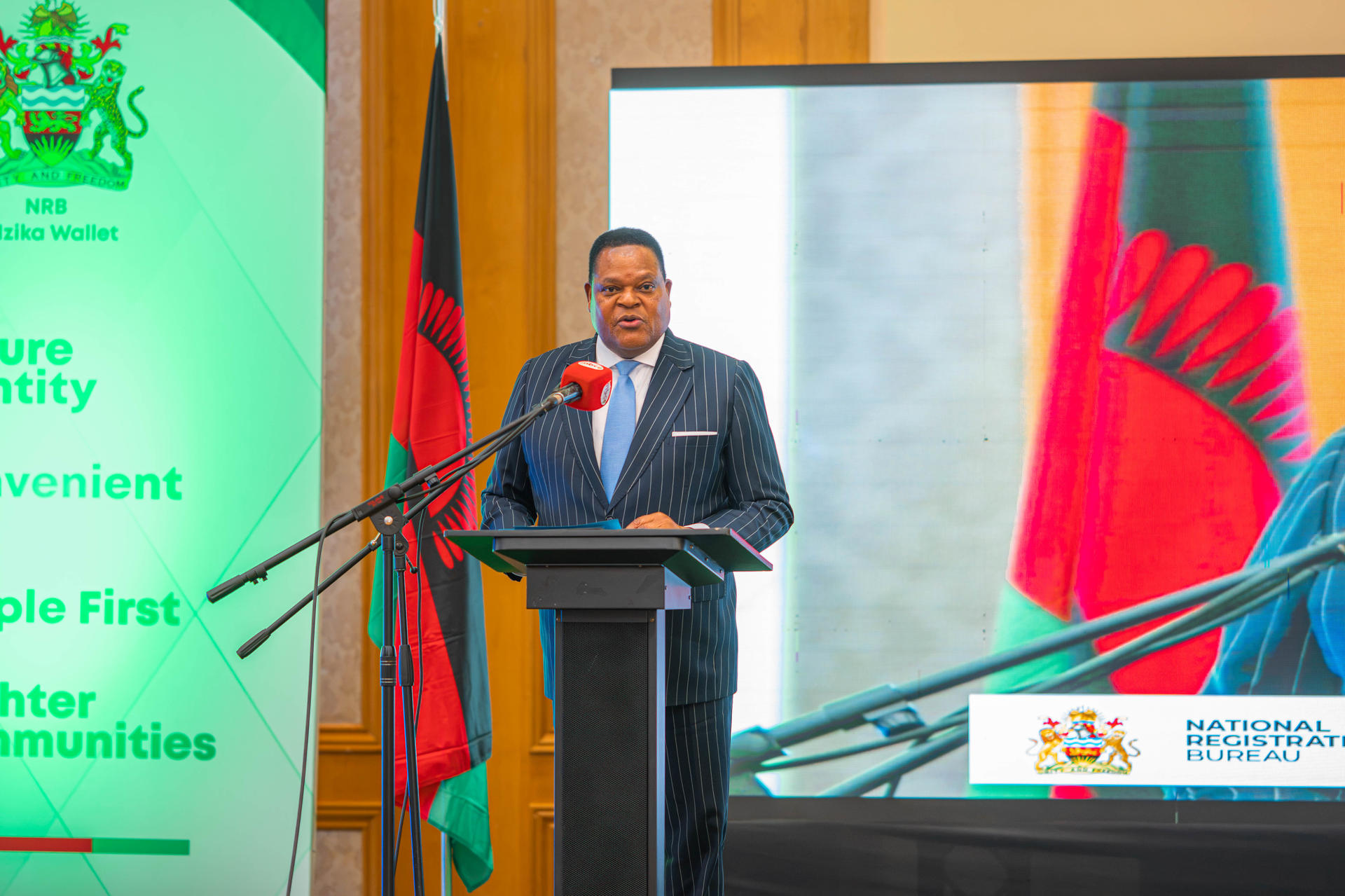 Photograph of a man in a suit speaking at a podium at a conference; flags and a screen behind.