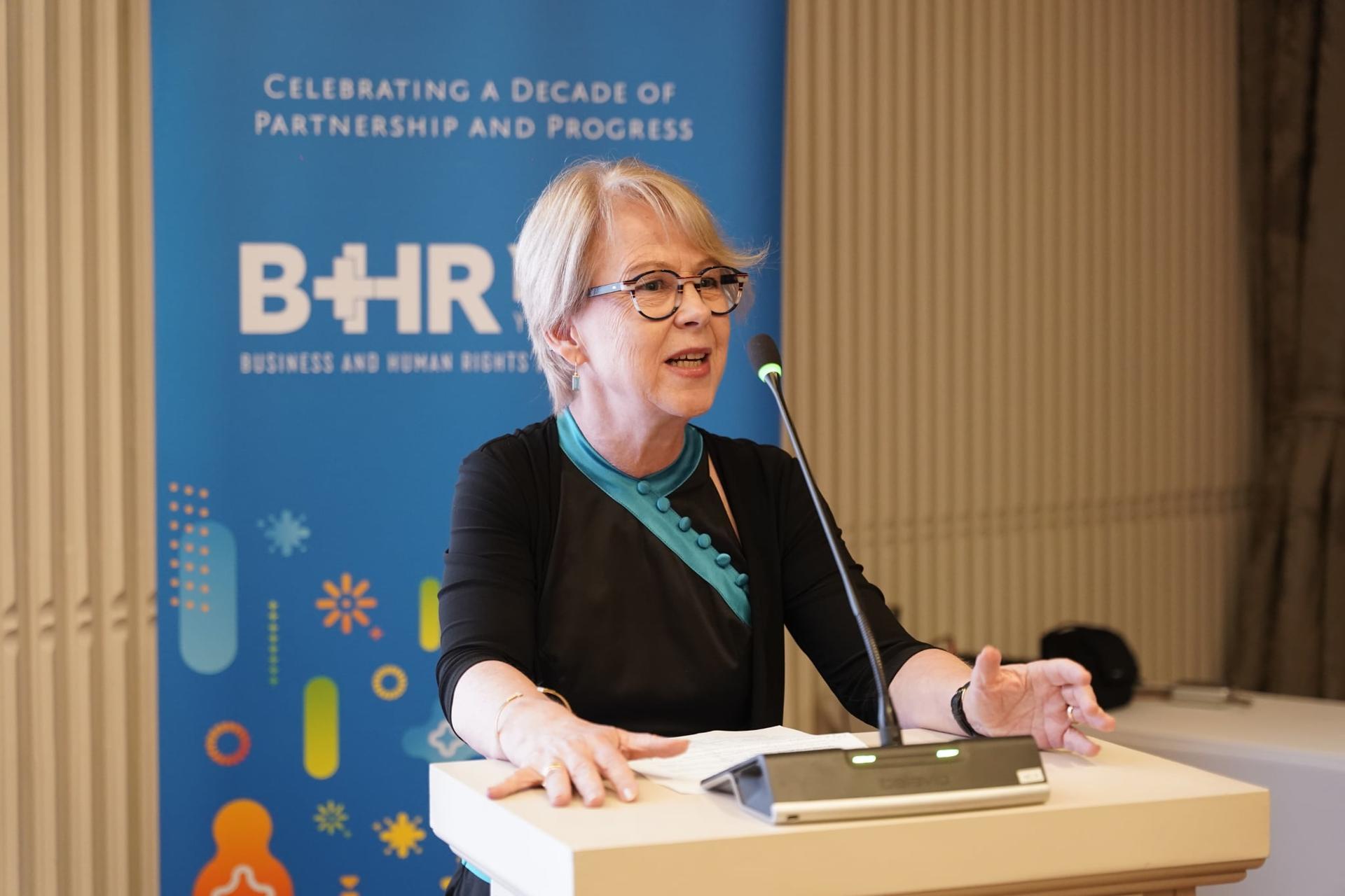 Older woman with glasses at podium, turquoise scarf, speaking; blue BHR banner behind.