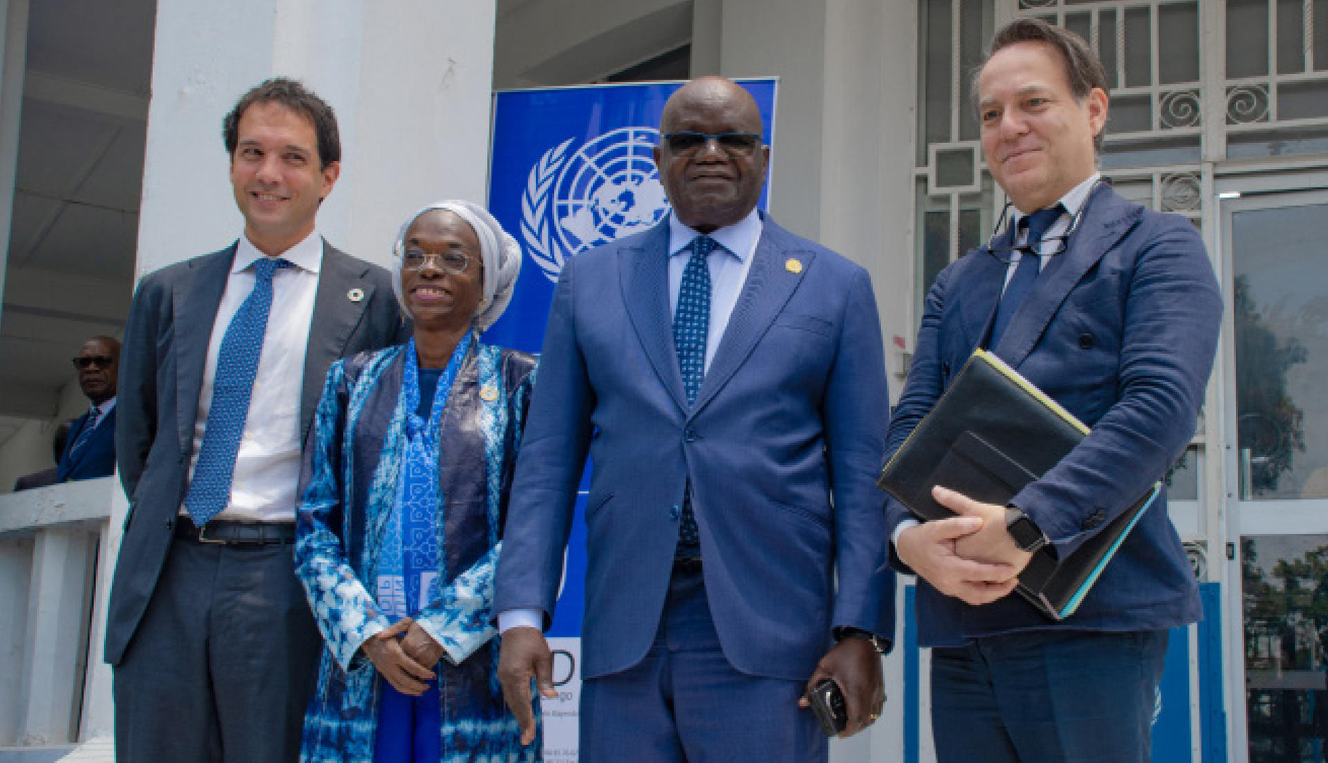 Four people in suits pose outside a building; a woman wearing a blue scarf, two men, and a man with a folder.