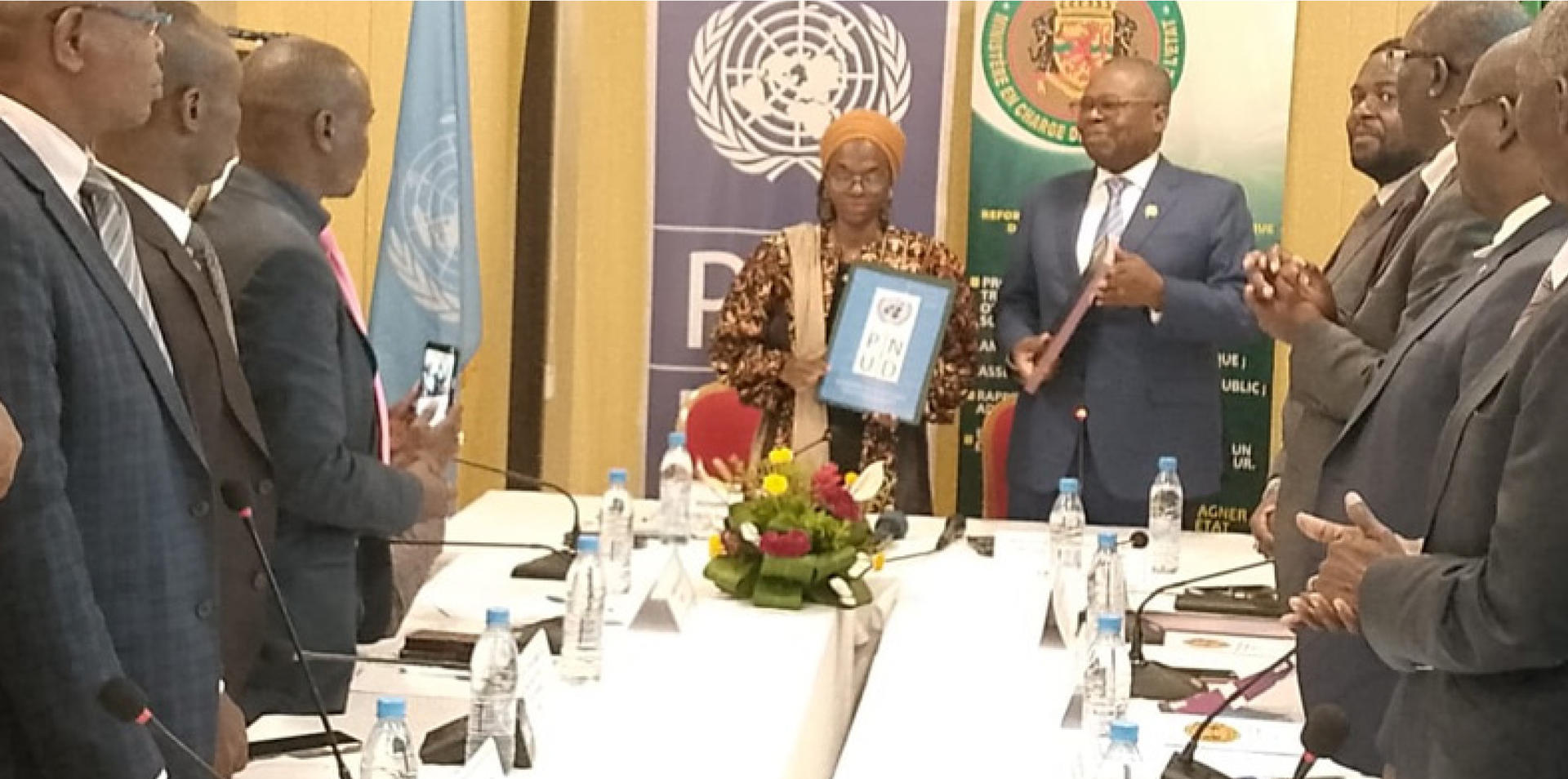Officials in suits present a framed certificate; flags and a flower centerpiece on the table.